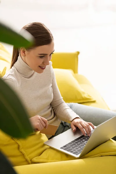 Selective focus of smiling girl using laptop and holding credit card on couch — Stock Photo