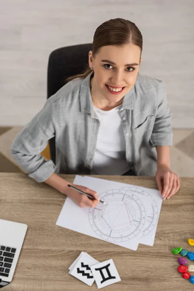 Hermoso astrólogo sonriente dibujando carta natal por cartas y piedras con signos del zodiaco en la mesa - foto de stock