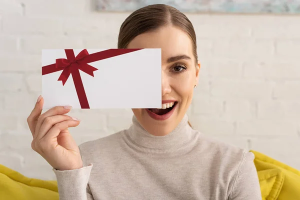 Atractiva chica salida cubriendo la cara con tarjeta de regalo y mirando a la cámara - foto de stock