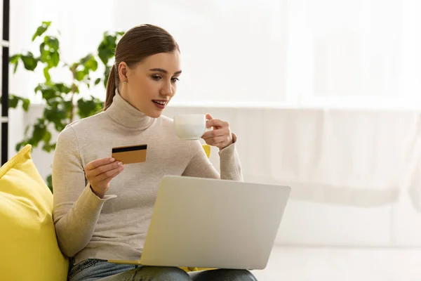 Приваблива молода жінка з ноутбуком і кредитною карткою п'є каву на дивані — стокове фото