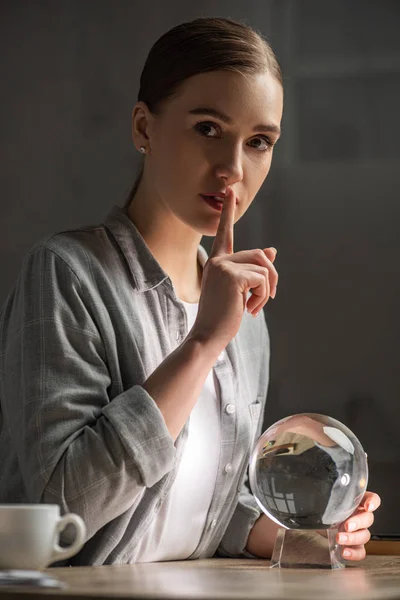 Attractive astrologer showing silence gesture and holding crystal ball on table — Stock Photo