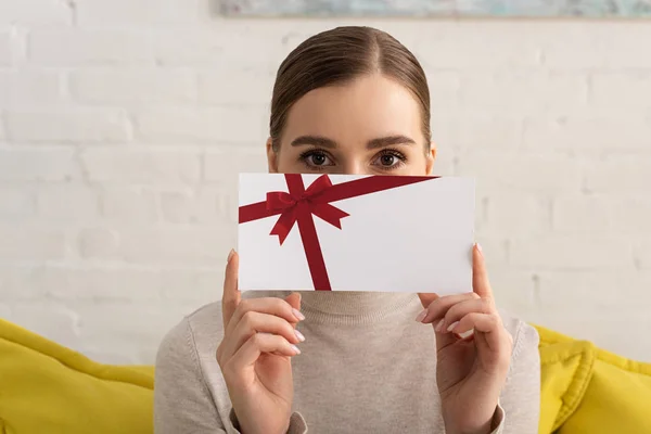 Mujer joven oscura cara con tarjeta de regalo y mirando a la cámara en el sofá - foto de stock