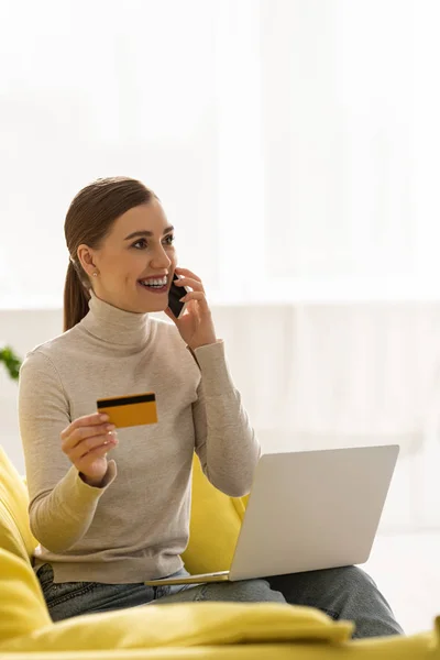 Femme souriante avec carte de crédit et ordinateur portable parlant sur smartphone sur le canapé — Photo de stock