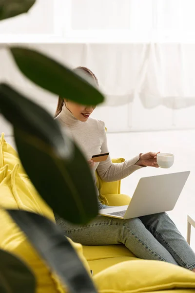 Selective focus of smiling girl holding credit card and coffee while using laptop on sofa — Stock Photo