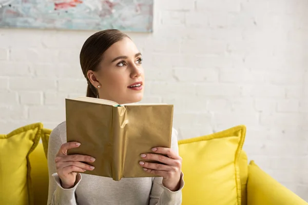 Задумчивая девушка держит книгу и смотрит на диван дома — стоковое фото