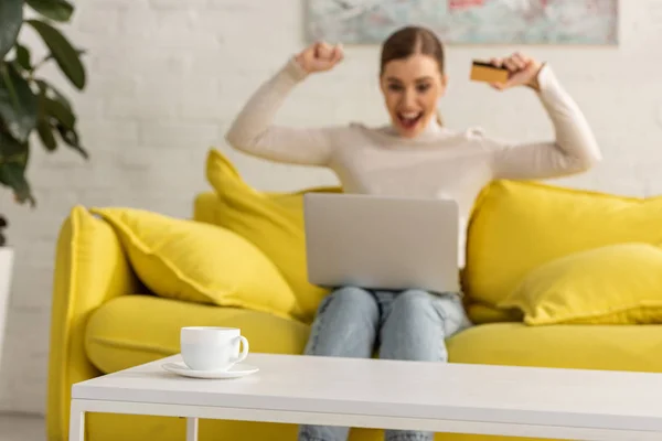 Enfoque selectivo de café en la mesa y mujer emocionada con tarjeta de crédito y portátil en el sofá en el fondo - foto de stock
