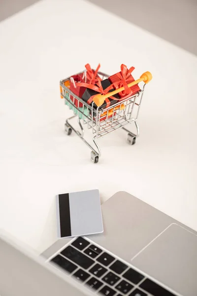 Laptop with credit card and toy gifts in cart on table — Stock Photo