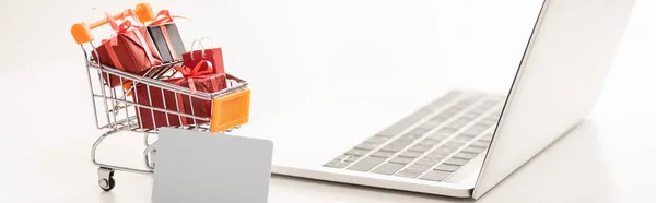Carrinho de brinquedo com caixas de presente ao lado de laptop e cartão de crédito na superfície branca, tiro panorâmico — Fotografia de Stock