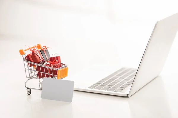Toy cart with gift boxes beside credit card and laptop on white surface — Stock Photo