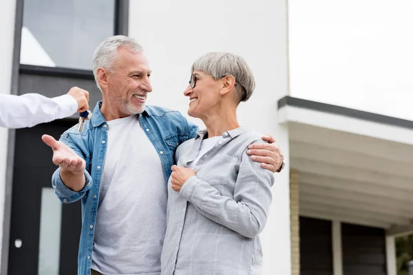 Cropped view of broker giving keys of new house to smiling mature man and woman — Stock Photo
