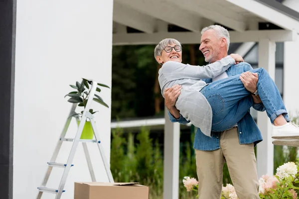 Mature man holding smiling woman in glasses near new house — Stock Photo