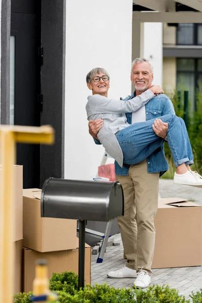 Mature man holding smiling woman in glasses near new house — Stock Photo