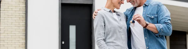 Panoramic shot of mature man holding keys of new house and hugging smiling woman — Stock Photo