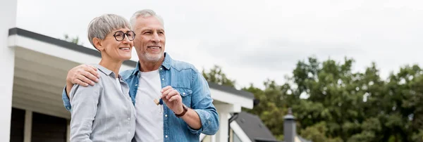 Panoramic shot of mature man holding keys of new house and hugging smiling woman — Stock Photo