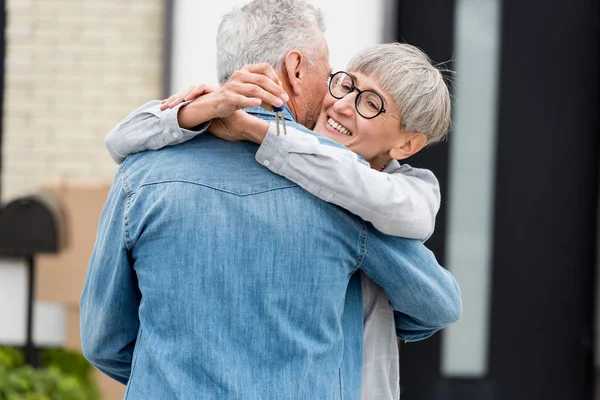 Mujer sonriente sosteniendo las llaves de la nueva casa y abrazando al hombre maduro - foto de stock