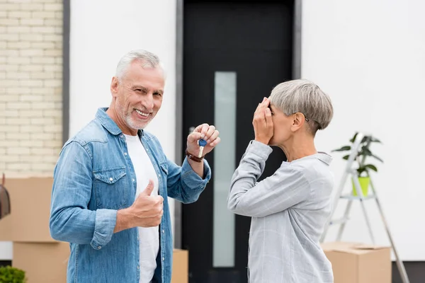 Älterer Mann hält Schlüssel für neues Haus und zeigt Gleichgesinnte und Frau verschleiert Gesicht — Stockfoto