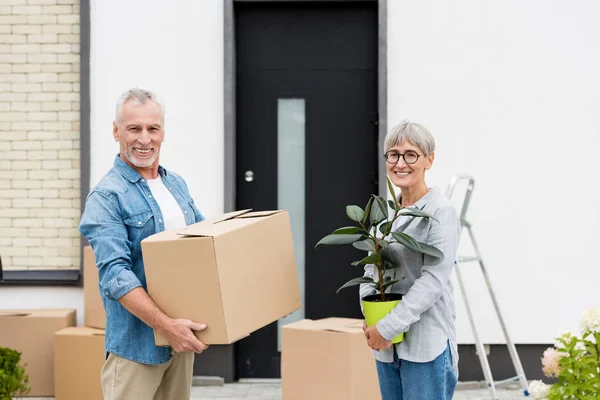 Älterer Mann hält Schachtel und lächelnde Frau hält Pflanze in der Nähe des neuen Hauses — Stockfoto