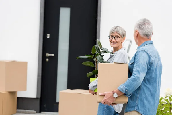 Hombre maduro sosteniendo caja y mujer sonriente sosteniendo planta cerca de casa nueva - foto de stock