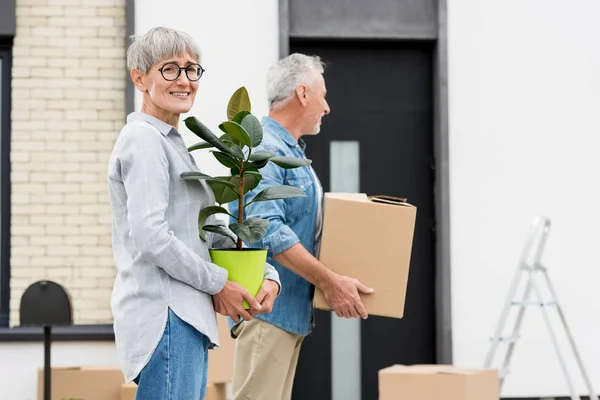Hombre maduro sosteniendo caja y mujer sosteniendo planta cerca de casa nueva - foto de stock