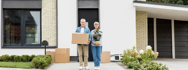 Tiro panorâmico de homem maduro segurando caixa e mulher segurando planta perto de nova casa — Fotografia de Stock