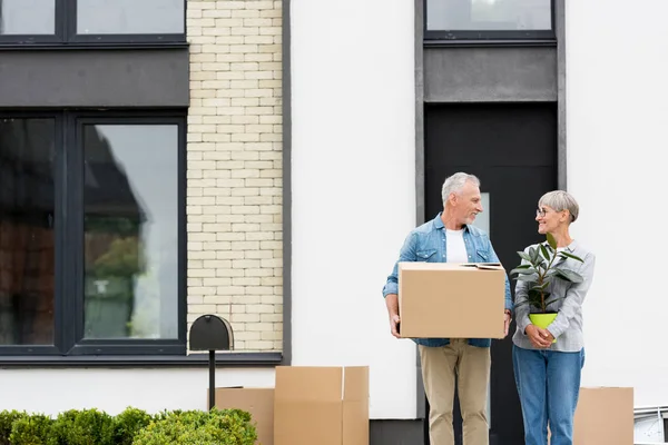 Lächelnder Mann mit Schachtel und Frau mit Pflanze in der Nähe des neuen Hauses — Stockfoto