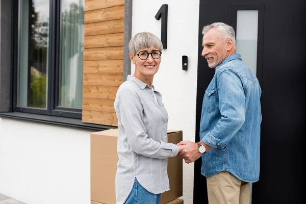 Mature man looking at smiling woman and holding hands with her new house — Stock Photo