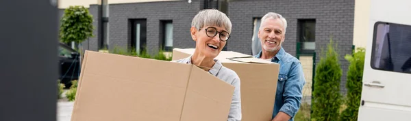 Panoramic shot of mature man and smiling woman bringing boxes to new house — Stock Photo