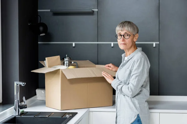 Зрелая женщина распаковывает коробку и смотрит на камеру в новом доме — стоковое фото