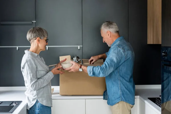 Вид сбоку зрелого мужчины и улыбающейся женщины, распаковывающих коробку в новом доме — стоковое фото