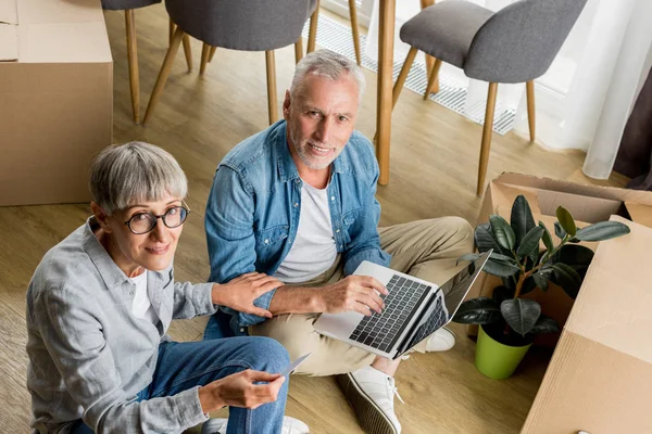 Vue grand angle de l'homme mature avec ordinateur portable et femme regardant la caméra dans une nouvelle maison — Photo de stock