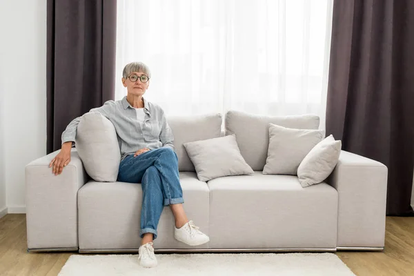 Mature femme dans lunettes assis sur canapé dans nouvelle maison — Photo de stock