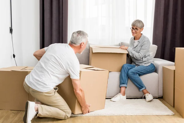 Mature man holding box and woman sitting on sofa in new house — Stock Photo