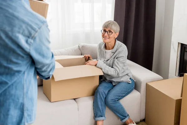 Cropped view of mature man holding box and smiling woman sitting on sofa in new house — Stock Photo