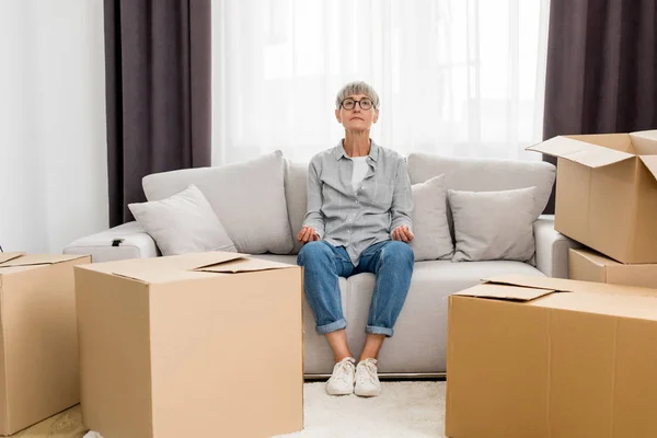 Mature woman sitting on sofa and meditating in new house — Stock Photo