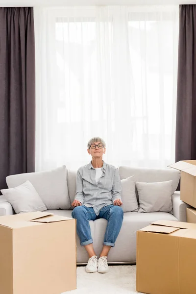 Mature woman with closed eyes sitting on sofa and meditating in new house — Stock Photo
