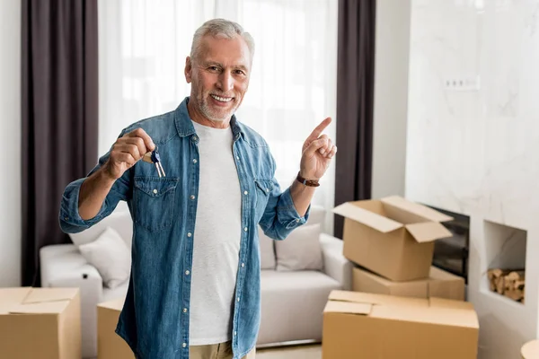 Smiling man holding keys and pointing with finger in new house — Stock Photo