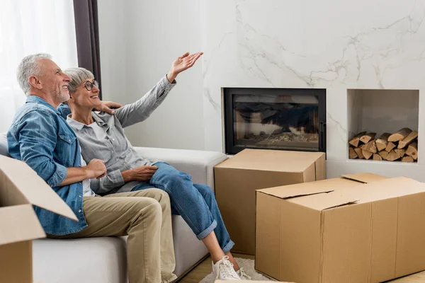 Mature man and woman sitting on sofa and pointing with hand in new house — Stock Photo