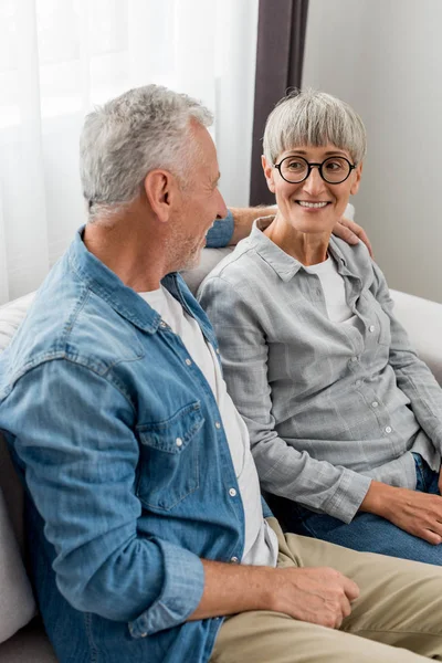 Зрелый мужчина обнимает улыбающуюся женщину и сидит на диване в новом доме — стоковое фото