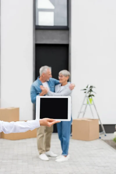 Foco seletivo de corretor segurando tablet digital e casal em pé perto de nova casa em segundo plano — Fotografia de Stock
