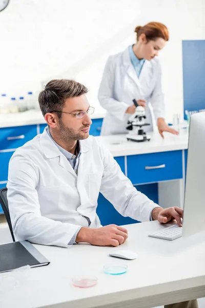 Selective focus of molecular nutritionist using computer and his colleague using microscope on background — Stock Photo