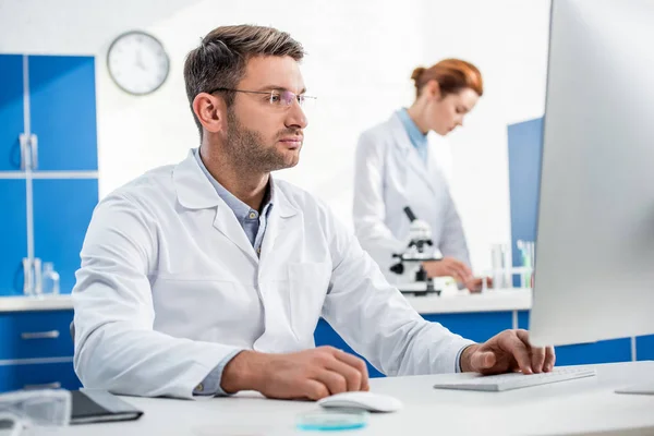 Selective focus of molecular nutritionist using computer and his colleague using microscope on background — Stock Photo