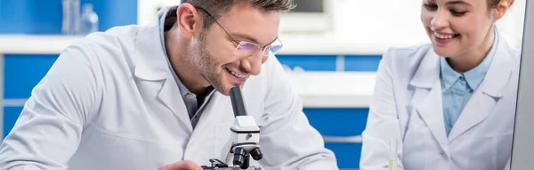 Panoramic shot of smiling molecular nutritionists using microscope in lab — Stock Photo