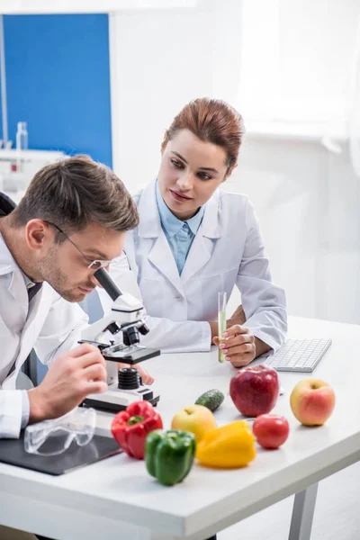 Nutricionista molecular usando microscopio y su colega sosteniendo tubo de ensayo - foto de stock