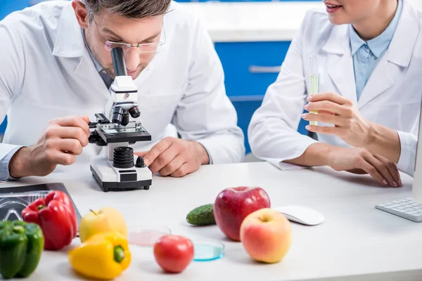 Visão cortada de nutricionista molecular segurando tubo de ensaio e seu colega usando microscópio — Fotografia de Stock