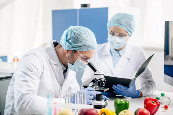 Carpeta de retención nutricionista molecular y mirando a colega con microscopio - foto de stock