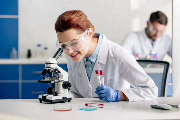 Foco seletivo de consultor genético sorrindo fazendo teste de dna e usando microscópio em laboratório — Fotografia de Stock