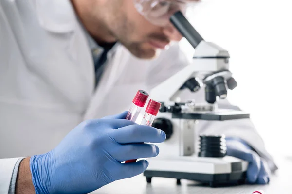 Vista recortada del consultor genético que sostiene tubos de ensayo y utiliza microscopio - foto de stock