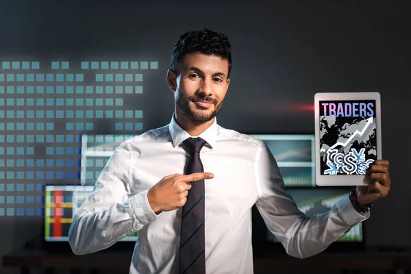 Happy bi-racial man pointing with finger at digital tablet with traders letters on screen — Stock Photo