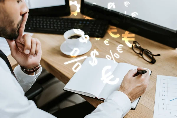 Vista recortada de comerciante bi-racial pensativo sentado en la mesa y la escritura en el cuaderno cerca de signos de dinero - foto de stock