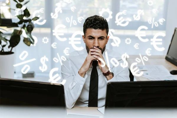 Задумчивый двурасовый торговец смотрит на компьютеры возле денежных знаков в офисе — стоковое фото